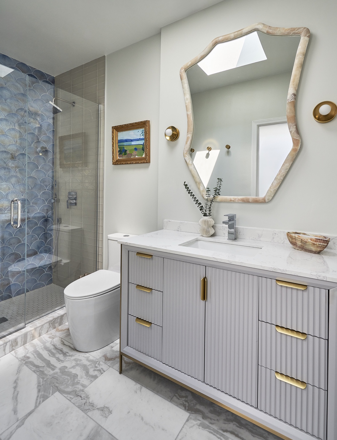 Blue scalloped shower tile, a ribbed vanity, and a sculptural mirror give this bathroom its character. Warm metal accents and artwork bring another layer of detail.
