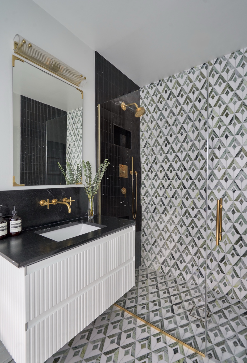 Green and white geometric tile extends across the floor and shower wall. Dark surfaces, a white ribbed vanity, and warm metal details bring contrast to the composition.